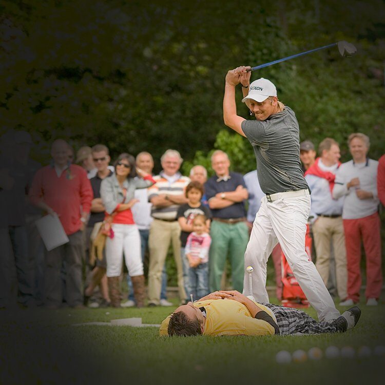 Mark Reynolds slaat tijdens een golf trickshow een bal van een tee die een vrijwilliger vasthoudt